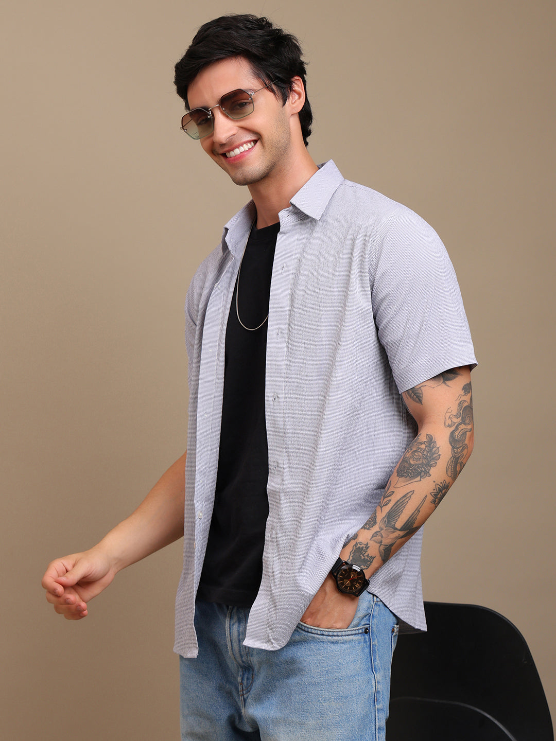 Man wearing sunglasses, a light gray shirt, black t-shirt, and jeans on a beige background