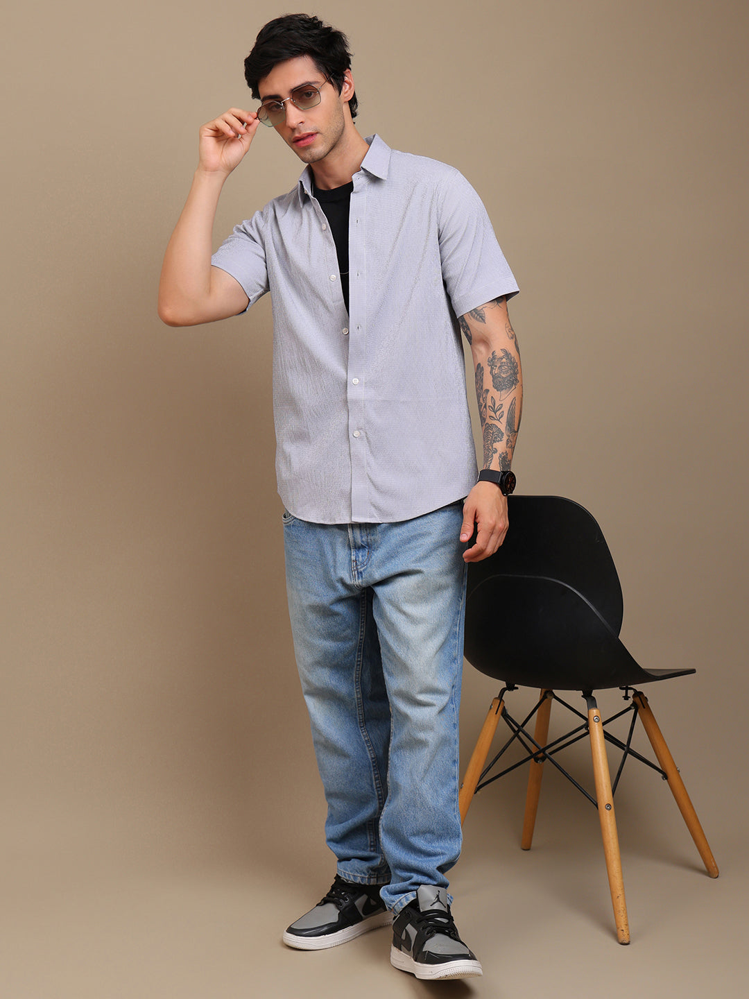 Man wearing a light gray shirt, black sunglasses, and blue jeans standing against a beige background with a black chair.